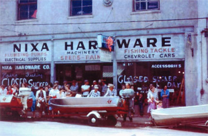 Historic Nixa Hardware storefront with three upstairs windows, boats, and a crowd outside windows marked Closed Sucker Day